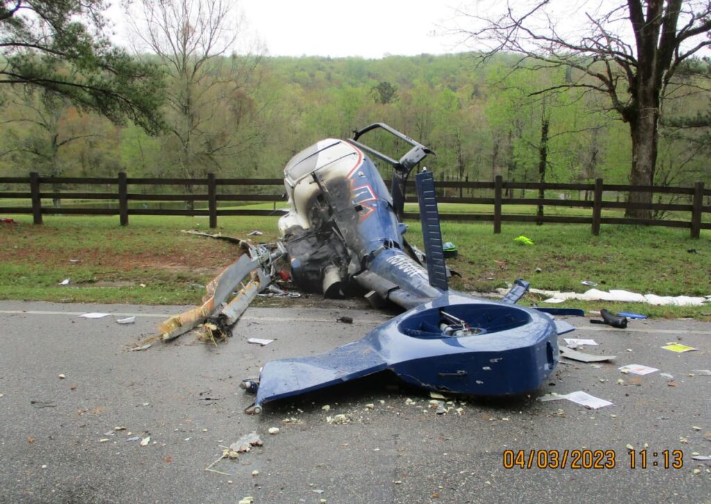 Frontal view of the crashed EC130 T2 helicopter on a road, with the tail rotor assembly detached and lying in the foreground, surrounded by scattered debris and a wooded area beyond.